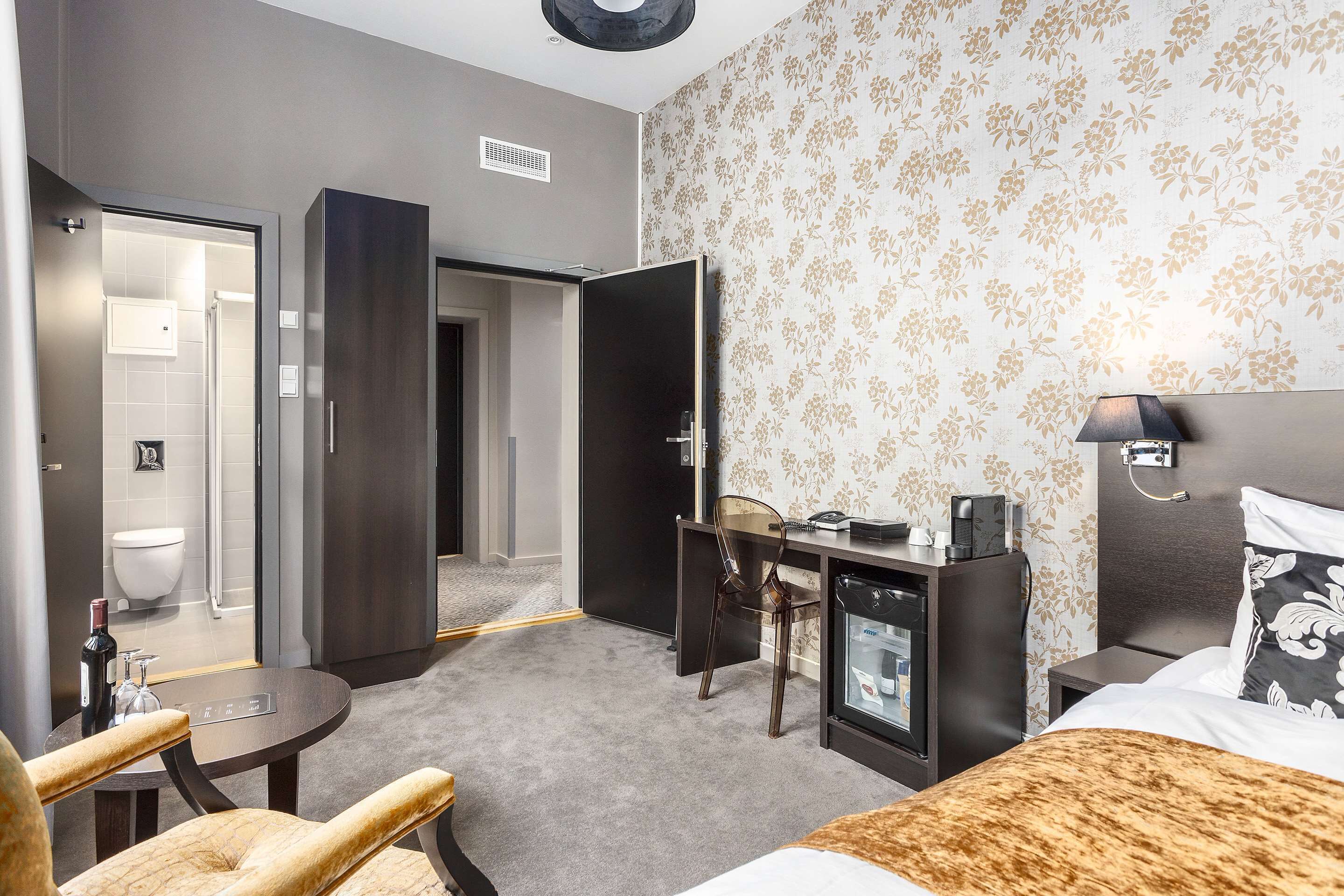 A hotel room with a bed, a desk, a chair, a minibar, and a bathroom visible through an open door.