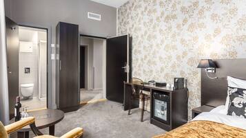 A hotel room with a bed, a desk, a chair, a minibar, and a bathroom visible through an open door.
