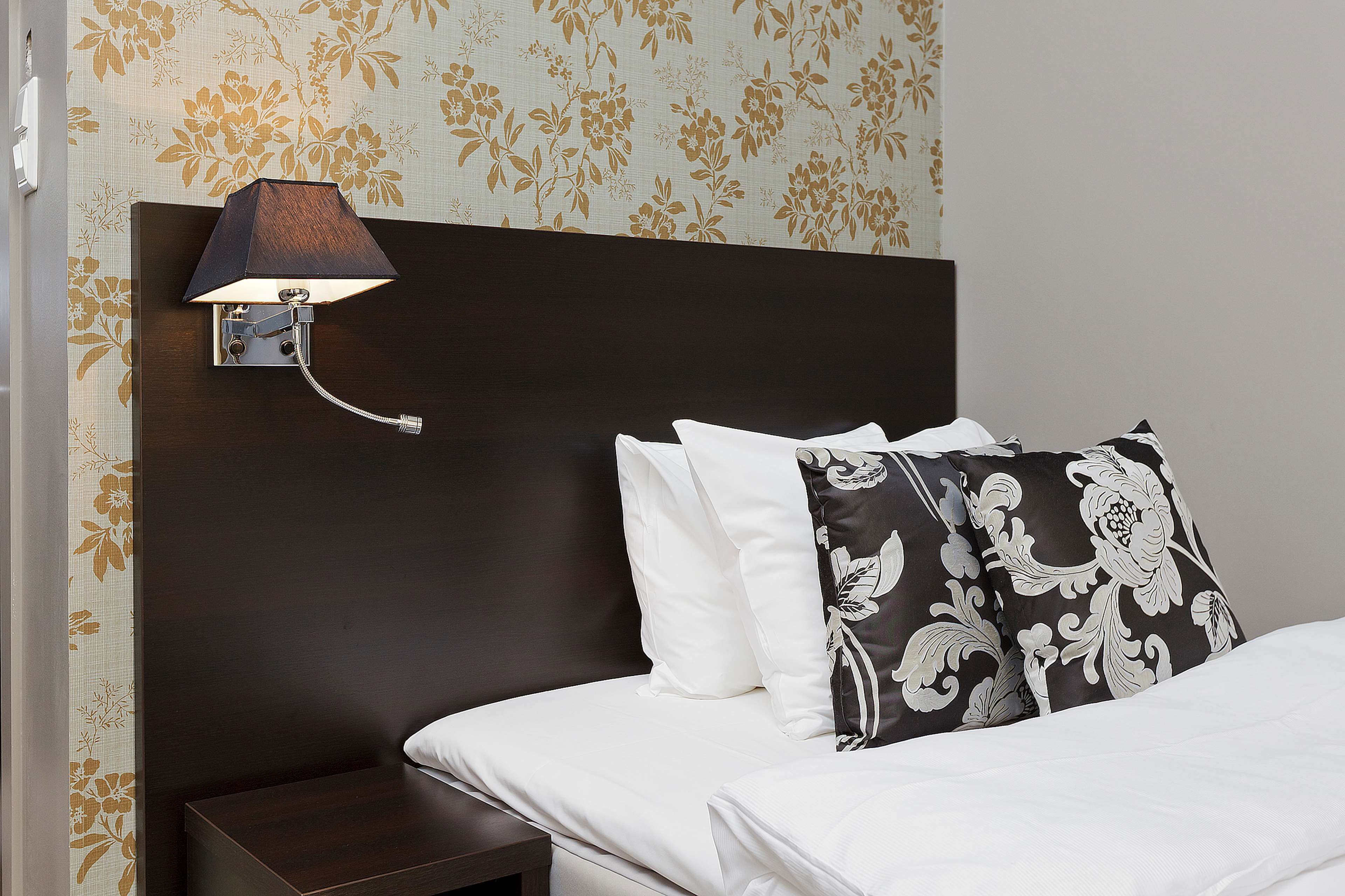 A neatly made bed with white bedding, a dark headboard, and a wall-mounted lamp.