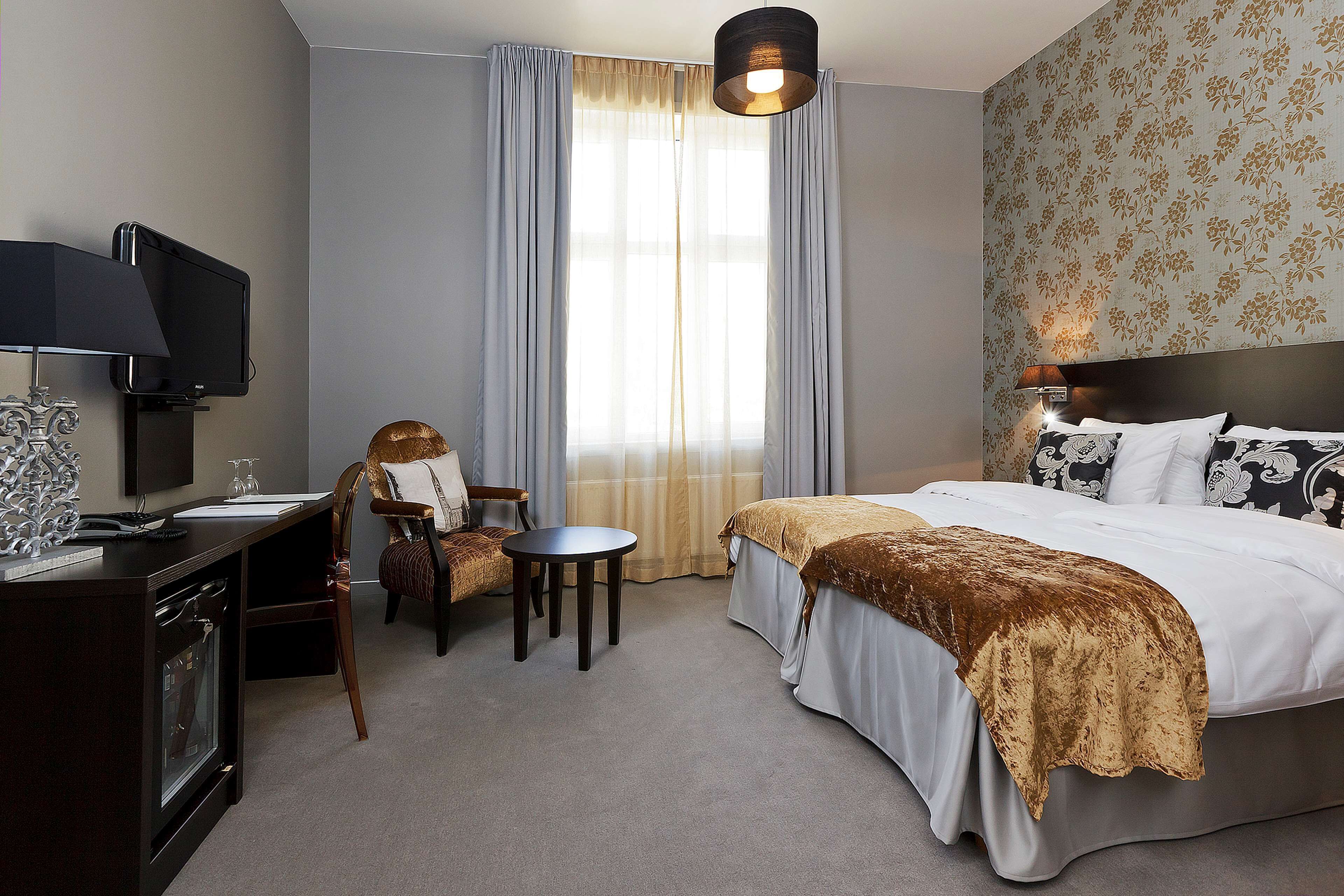 A hotel room with a bed, a desk with a TV, a chair, a small table, and a window with curtains.