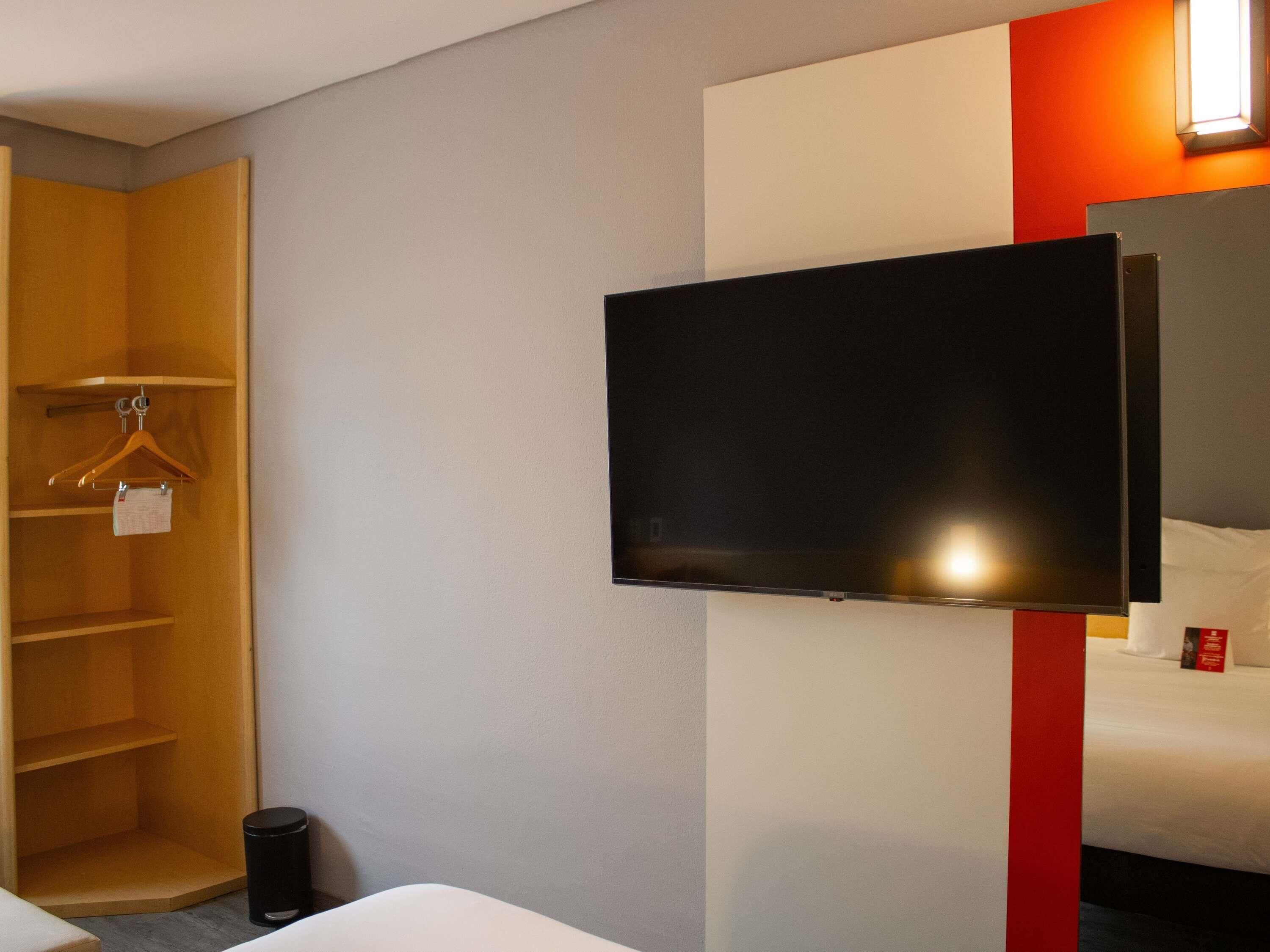Hotellihuone, jossa on sänky, seinälle asennettu televisio, vaatekaappi ja roskakori.
