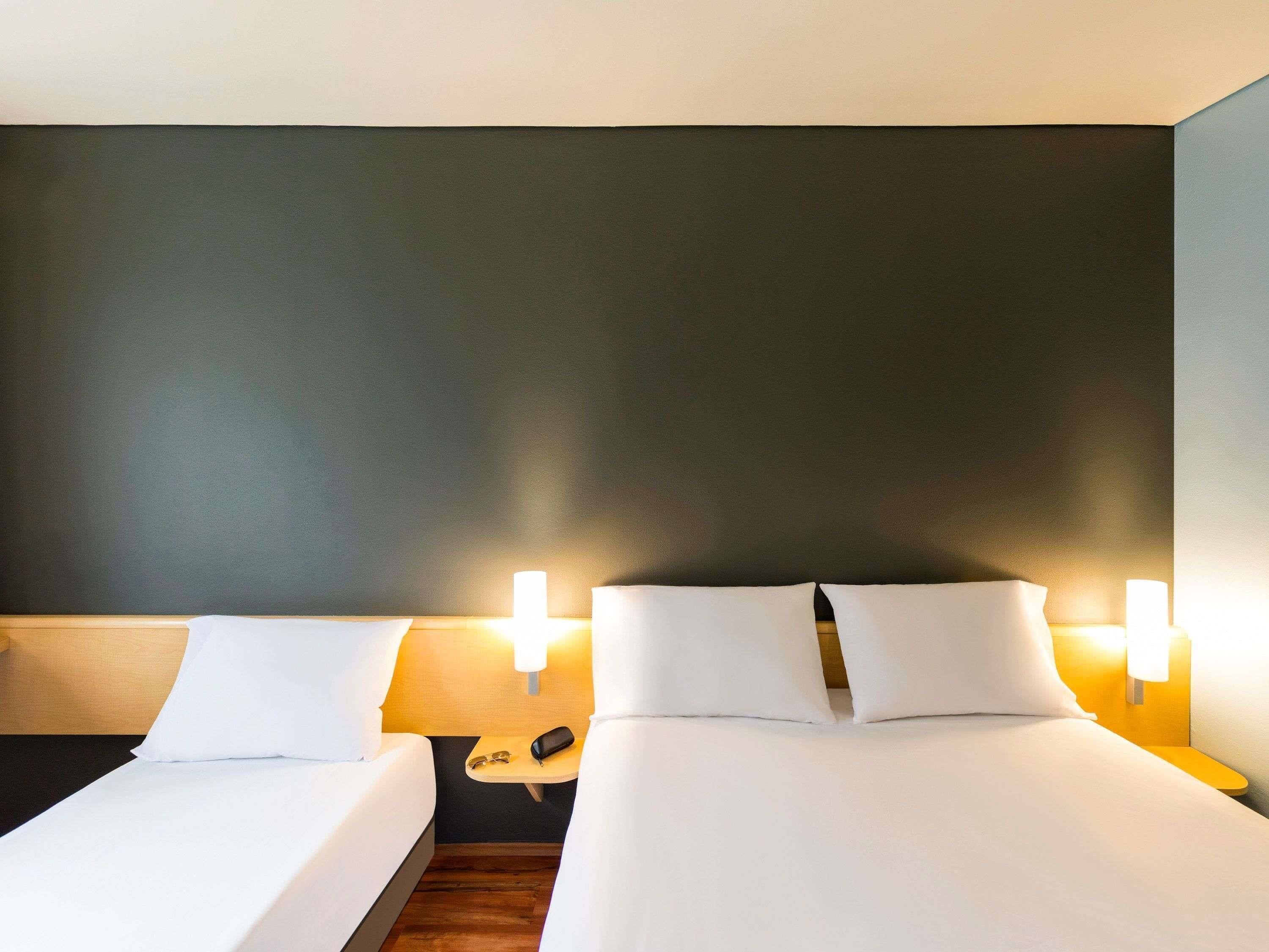 A hotel room with two beds, bedside lamps, a nightstand with a phone and glasses, and a dark-colored wall.