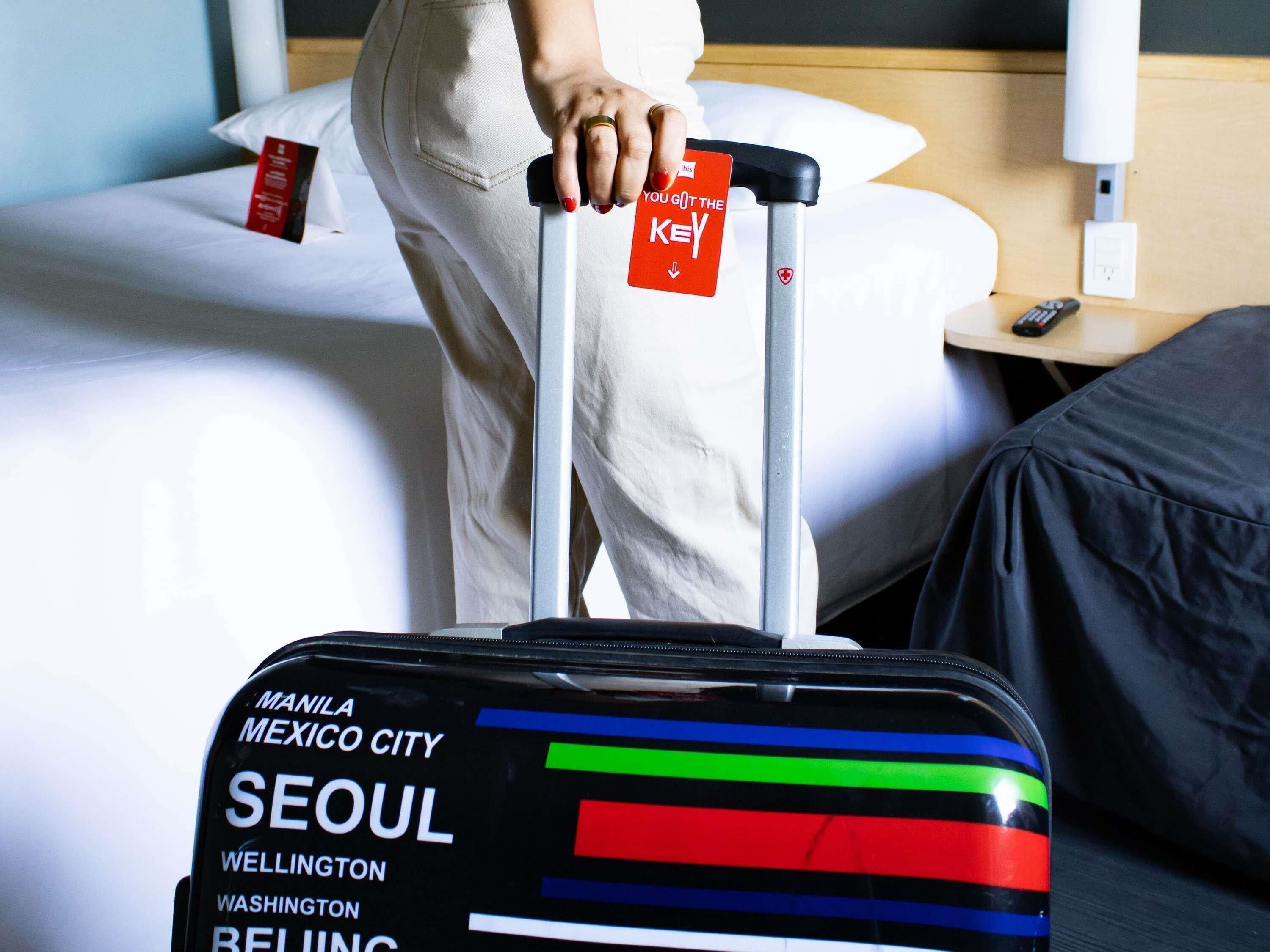 A person pulling a suitcase with a key card labeled 'YOU GOT THE KEY' and a hotel room with a bed, a nightstand, and a remote control.