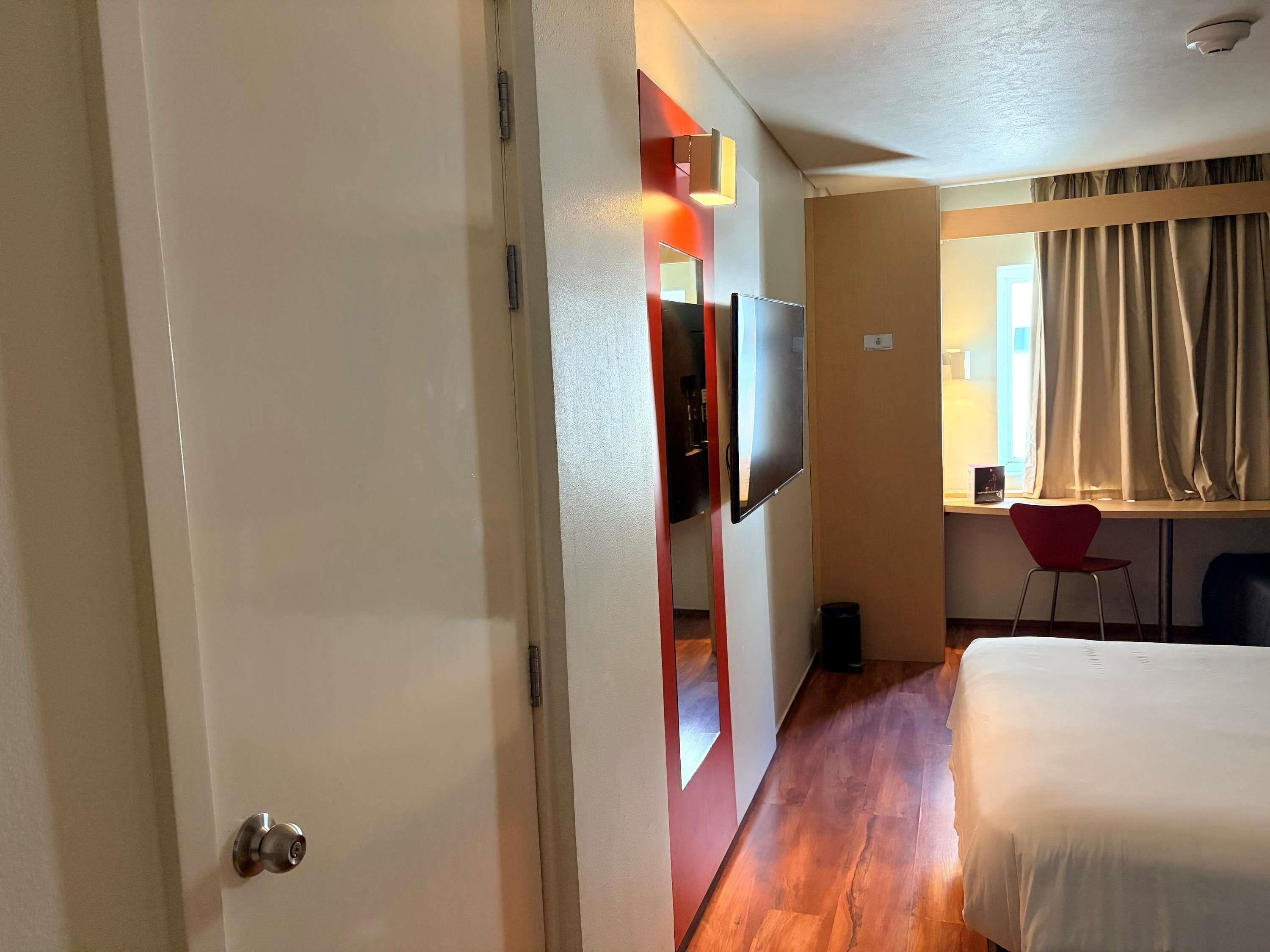 A hotel room with a bed, a TV mounted on the wall, a desk with a chair, and a window with curtains.