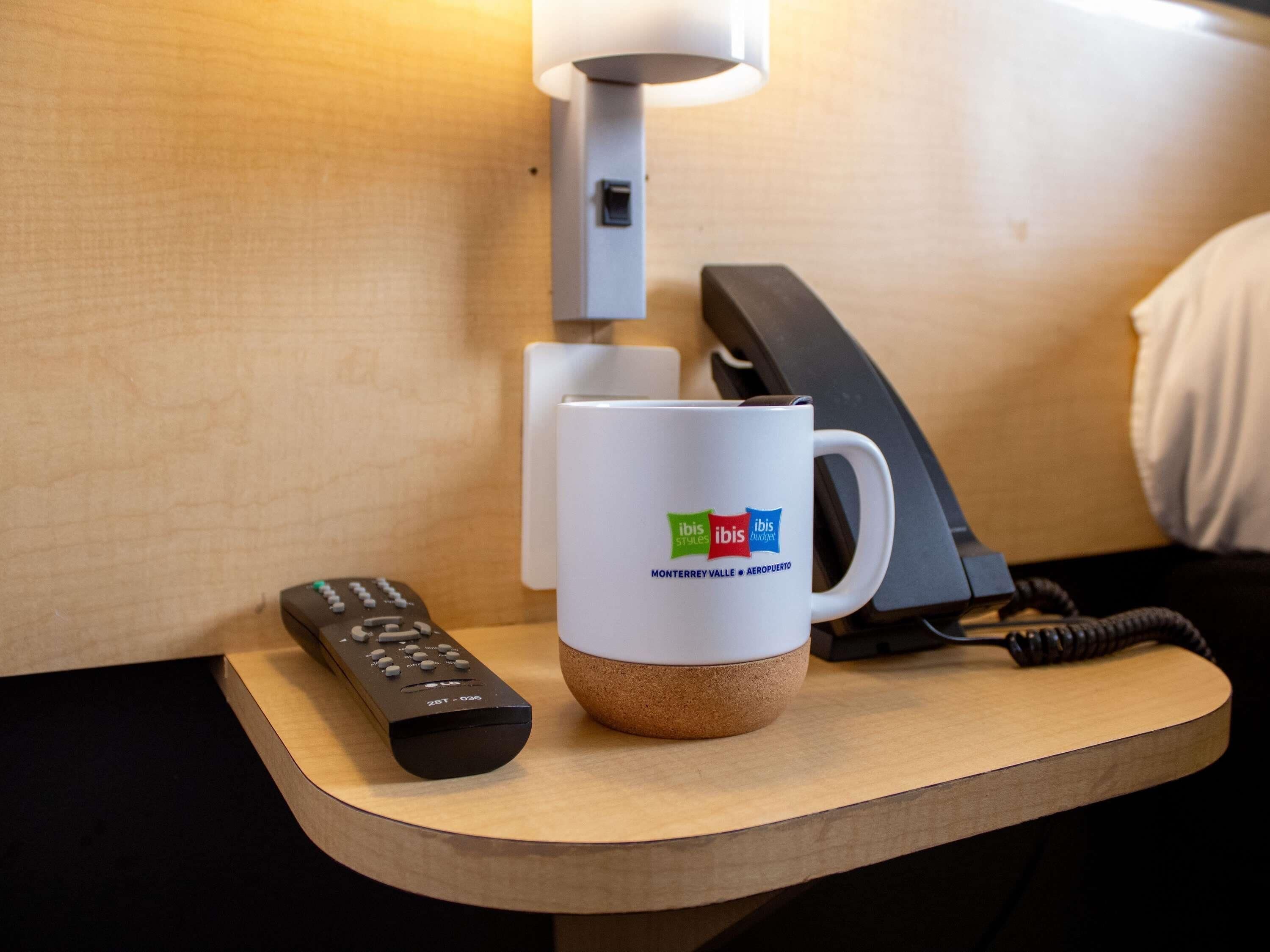 A hotel room with a wooden nightstand, a white mug with logos, a remote control, and a telephone.