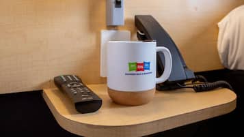 A hotel room with a wooden nightstand, a white mug with logos, a remote control, and a telephone.
