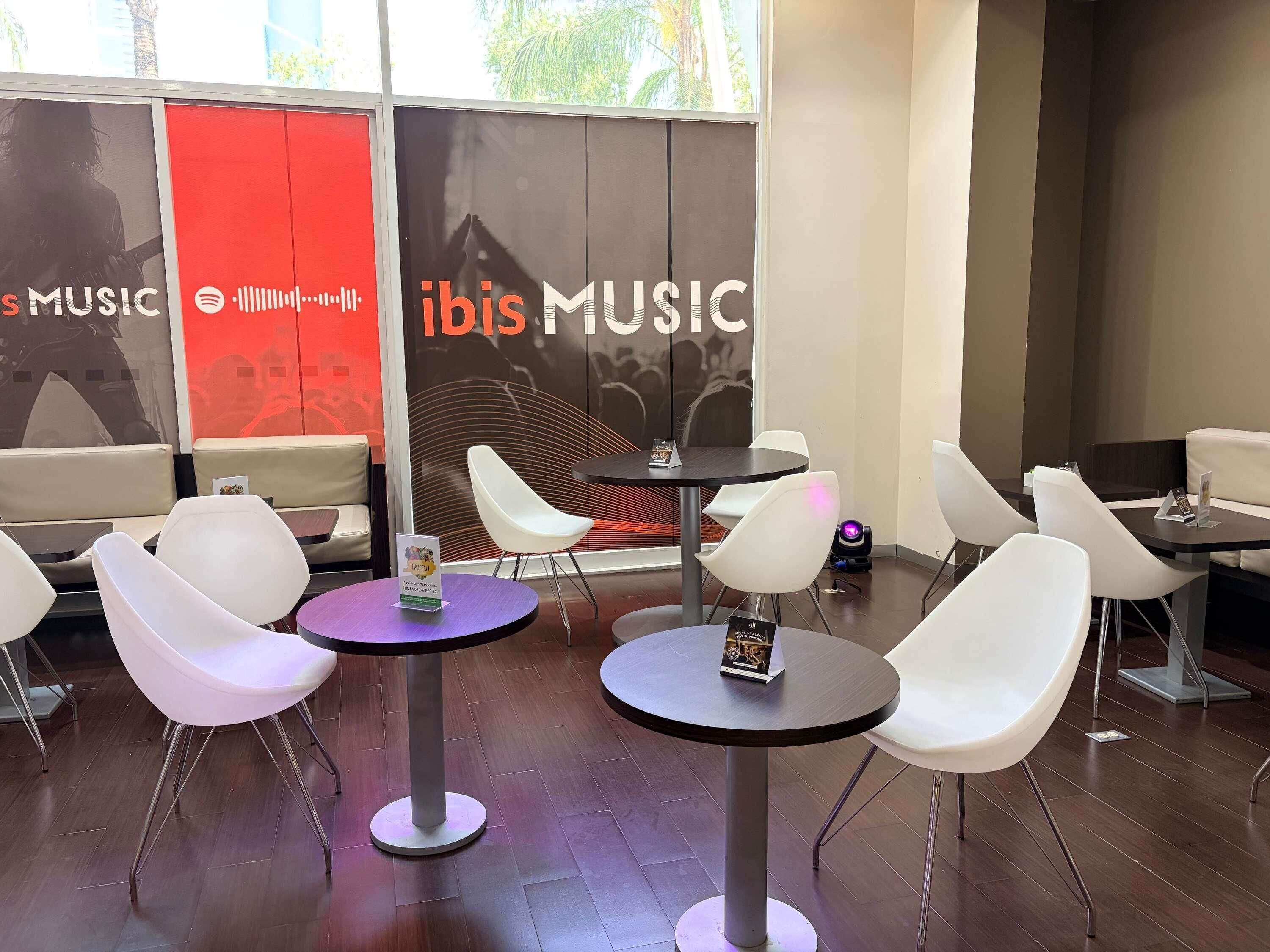 Un intérieur moderne avec des tables rondes et des chaises blanches, une grande fenêtre ornée du logo « ibis MUSIC » et un mur d’accent rouge.