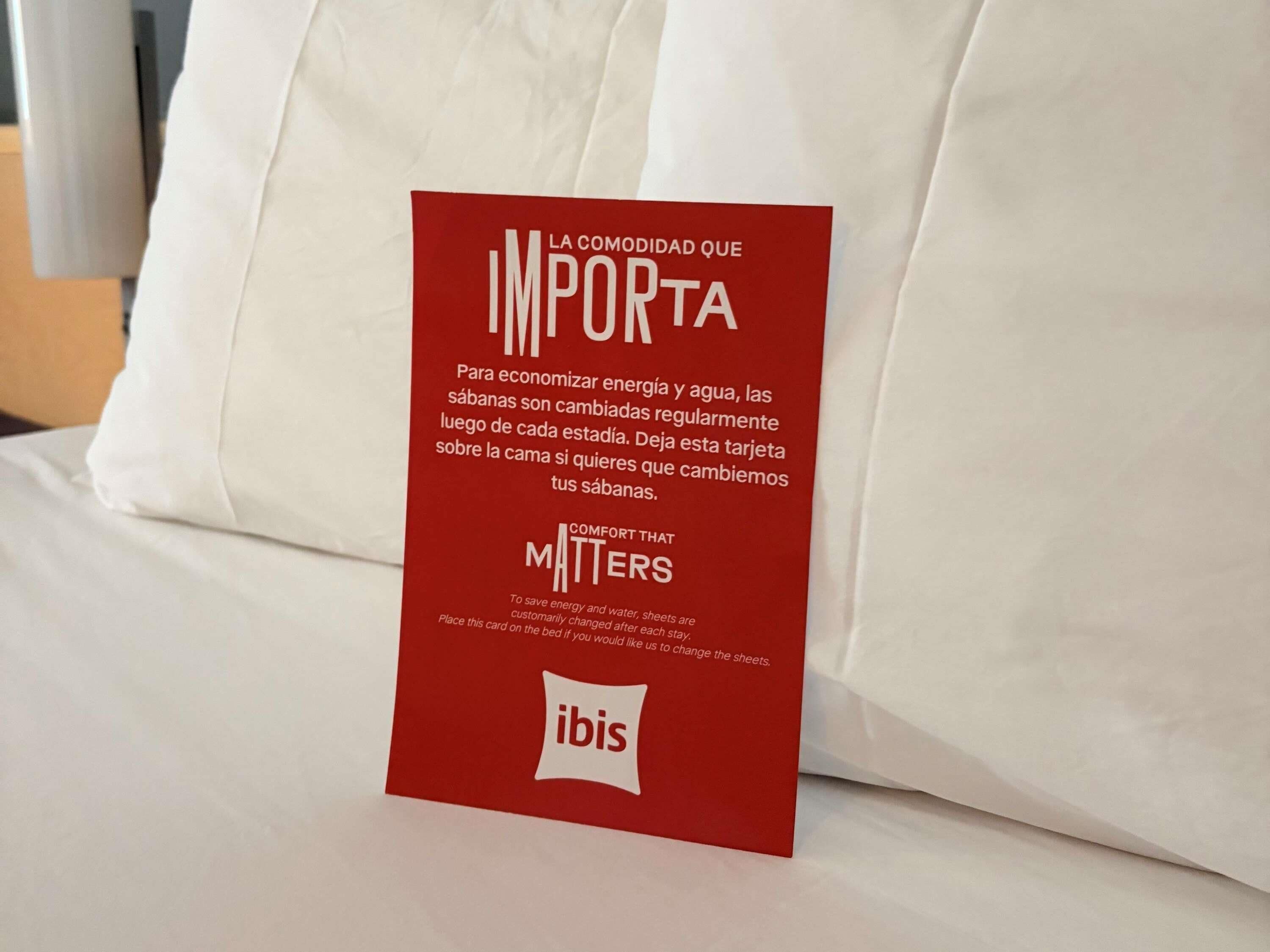 A red card on a bed with white bedding, containing text in Spanish and English about energy and water conservation, and the ibis hotel logo.