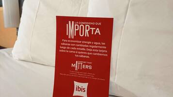 A red card on a bed with white bedding, containing text in Spanish and English about energy and water conservation, and the ibis hotel logo.