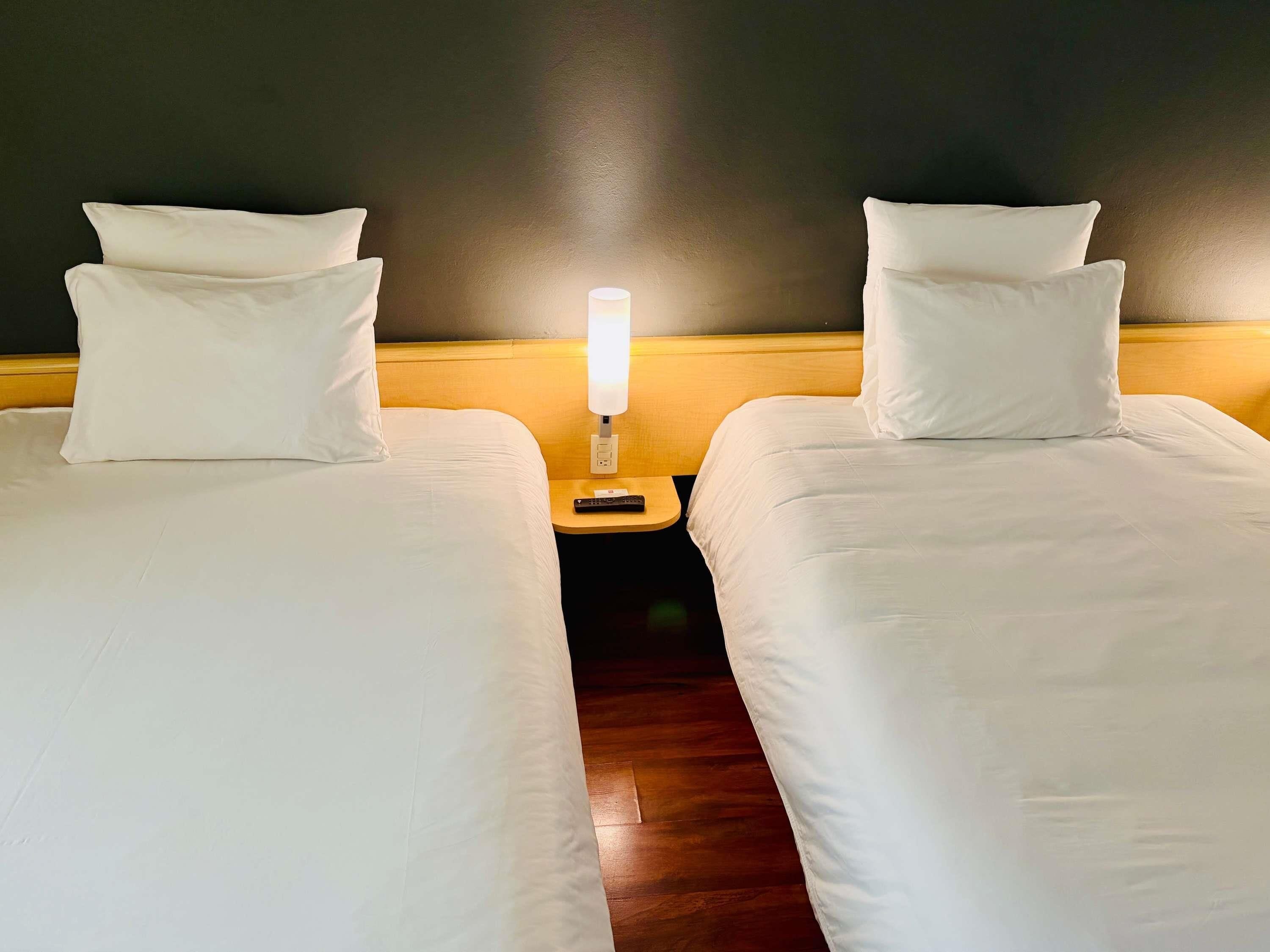 A hotel room with two single beds, a nightstand with a lamp, and a wooden headboard.