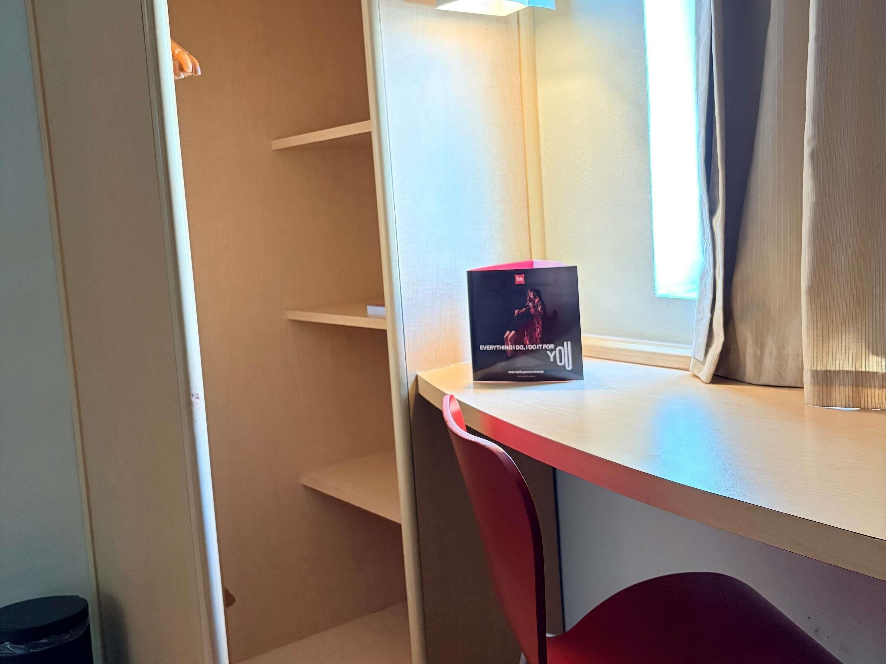 A hotel room with a desk, a chair, a window with curtains, and a trash bin.