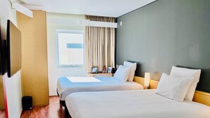 Habitación de hotel con dos camas, piso de madera, ventana con cortinas y mesita de noche con lámpara.