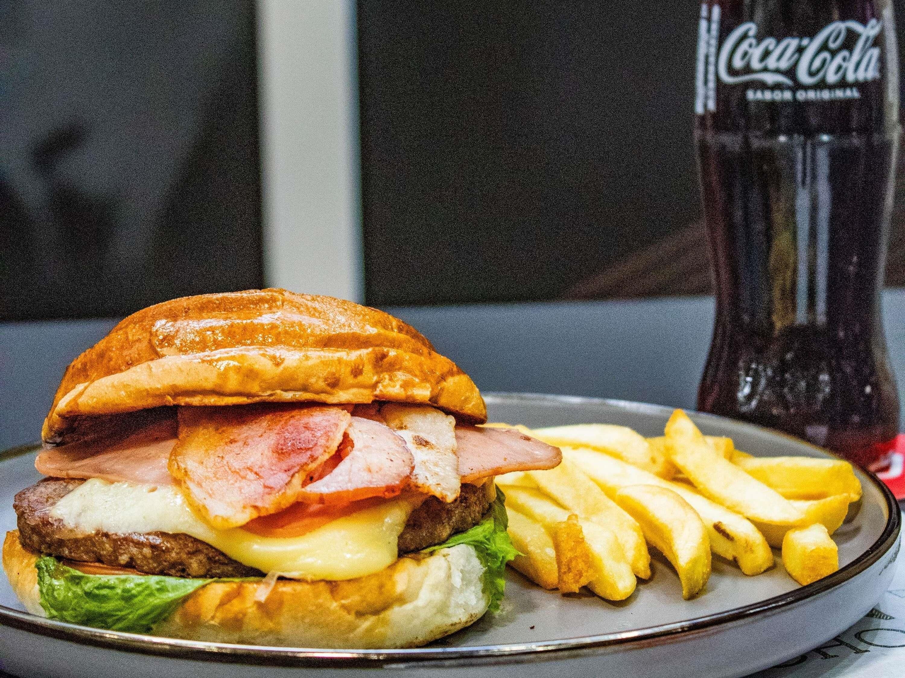 Ein Sandwich mit Speck, Käse und Salat auf einem Teller, dazu Pommes und eine Coca-Cola Flasche.