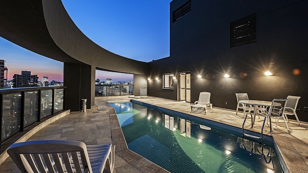 A rooftop pool area with a clear view of the city skyline at dusk, a pool with a tiled edge, and a seating area with chairs and a table. - SLAVIERO Moema (São Paulo)