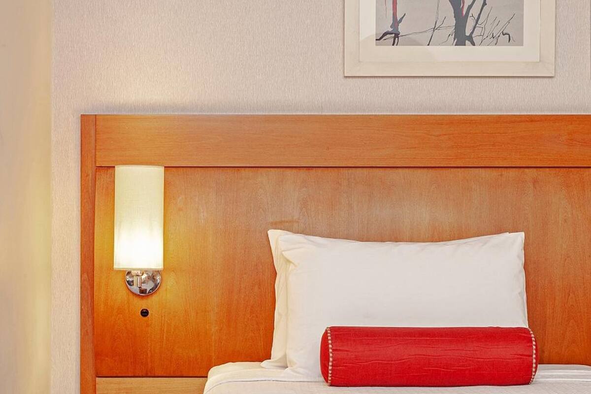 A hotel room with a wooden headboard, a bed with white and red bedding, a framed artwork on the wall, and a wall-mounted light fixture.