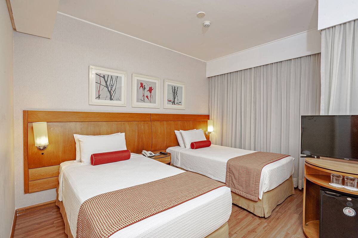 A hotel room with two beds, a TV, a minibar, and artwork on the wall.