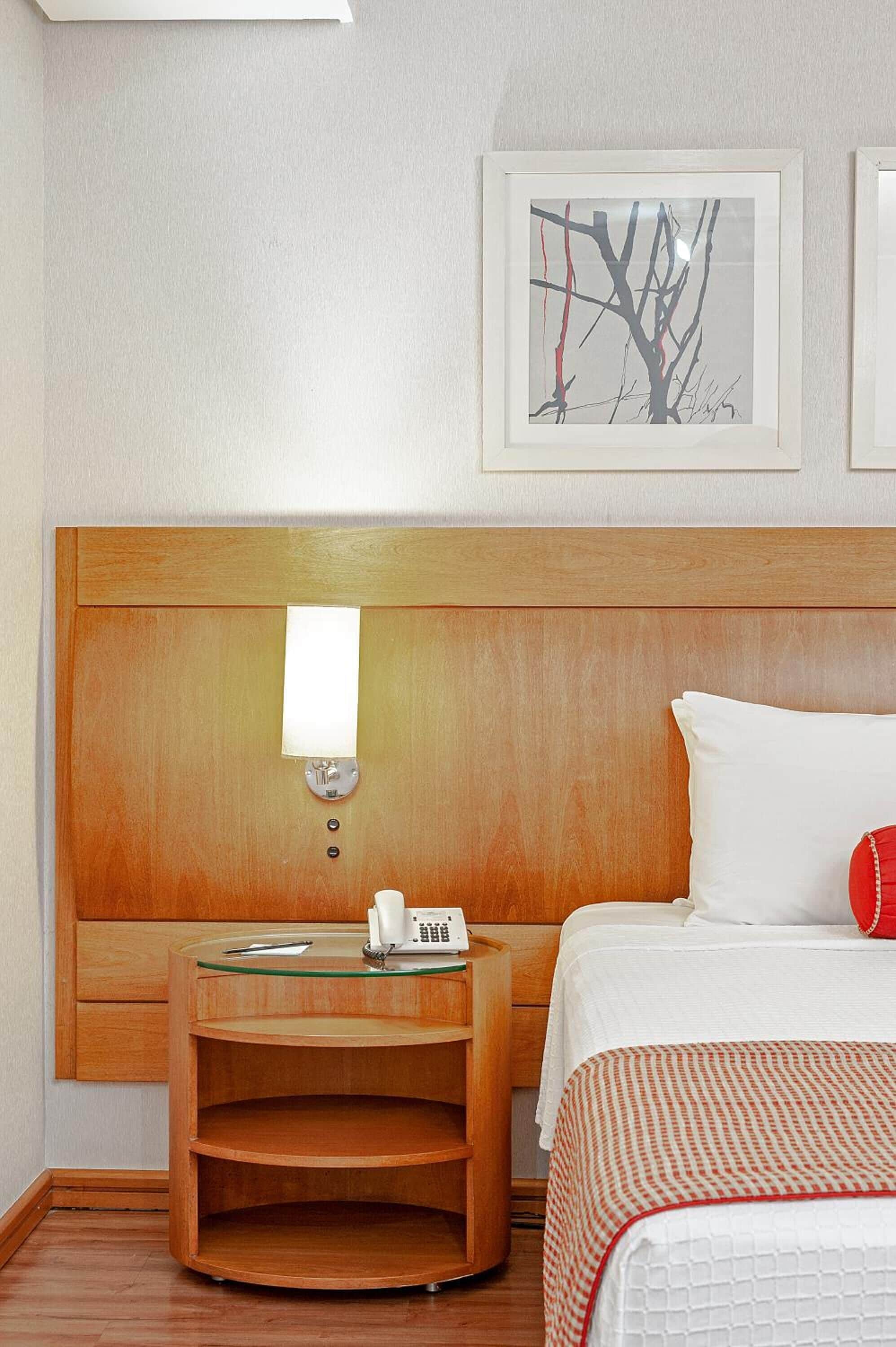 A hotel room with a wooden headboard, a bedside table with a lamp, a telephone, and a framed picture on the wall.