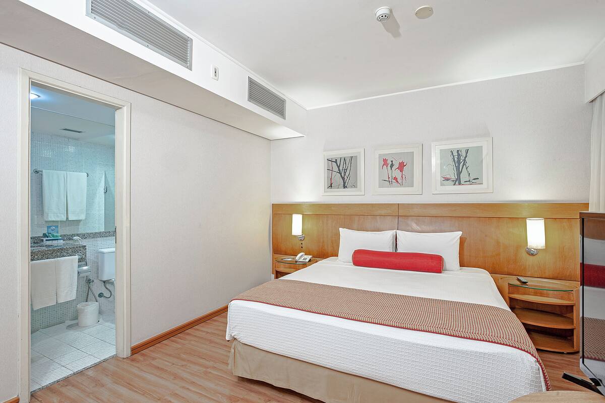 A hotel room with a large bed, a bedside table, a nightstand, a mirror, and a bathroom visible through an open door.
