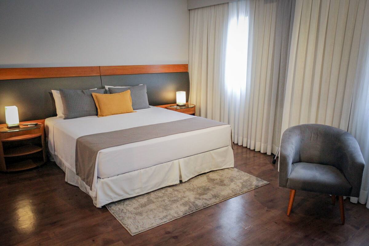 A hotel room with a large bed, two bedside tables, a chair, and a window with curtains.