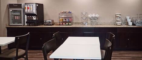 Una habitación pequeña y limpia con una mesa blanca y cuatro sillas, un refrigerador, una cafetera y un estante con diversos objetos.