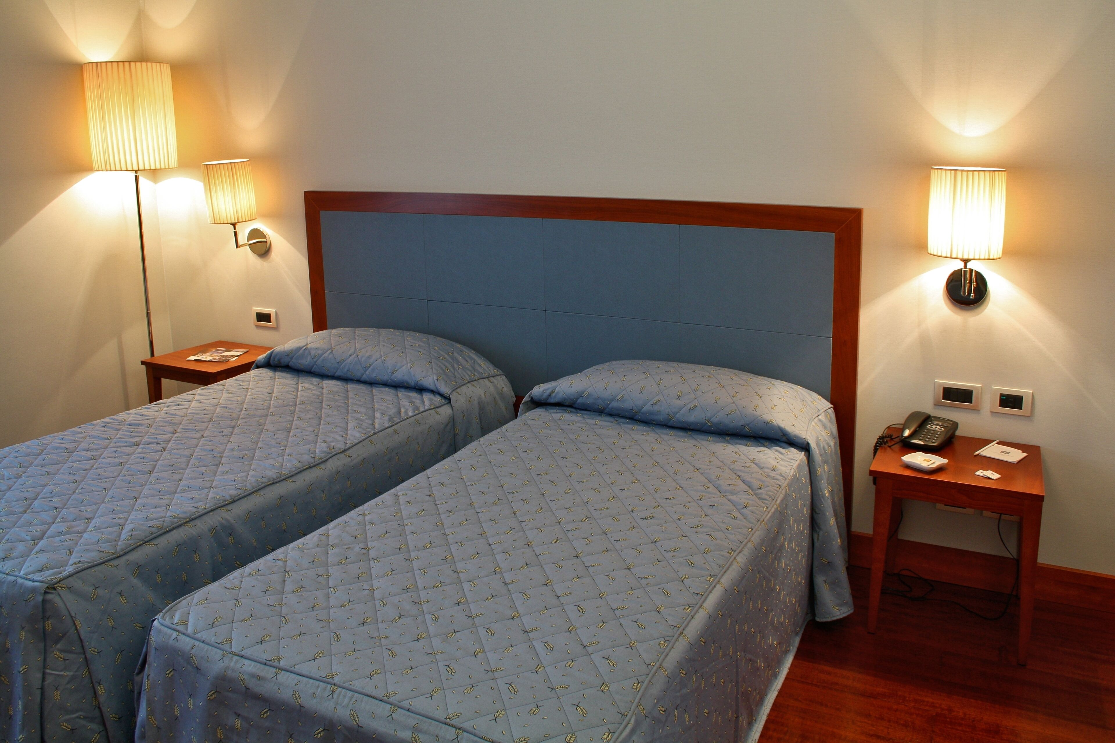 Ein Hotelzimmer mit zwei Einzelbetten, jeweils mit blauer, gesteppter Bettdecke und passenden Kissen. Es gibt Nachttische mit Lampen, ein Telefon und einen kleinen Tisch mit einem Buch und einem Notizblock.