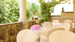 Un área de comedor al aire libre con manteles blancos y sillas, un arreglo floral rosa y vistas a la vegetación y una palmera.