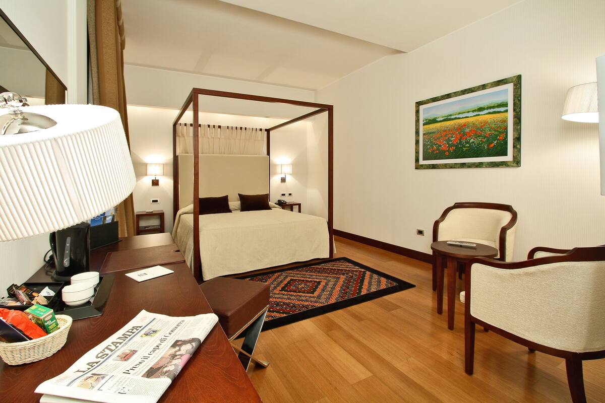 Une chambre d’hôtel avec un lit à baldaquin, un bureau avec une lampe, une chaise, une petite table et un tableau au mur.