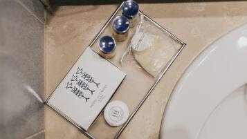 Un baño con pared de baldosas color beige, un inodoro blanco, y una pequeña bandeja con artículos de aseo personal, incluyendo una pastilla de jabón, una botella de loción y un pequeño recipiente.