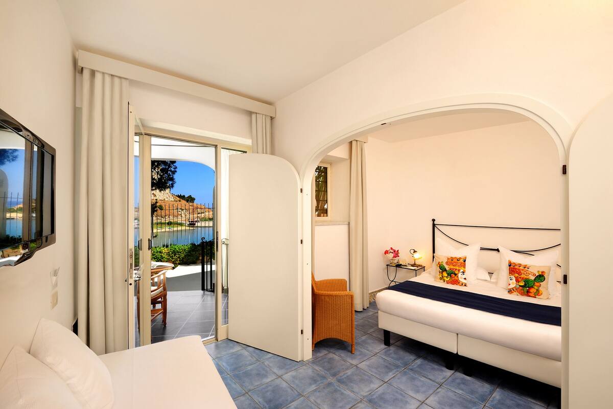 A bedroom with a bed, a sofa, a chair, and a view of a pool and greenery outside.