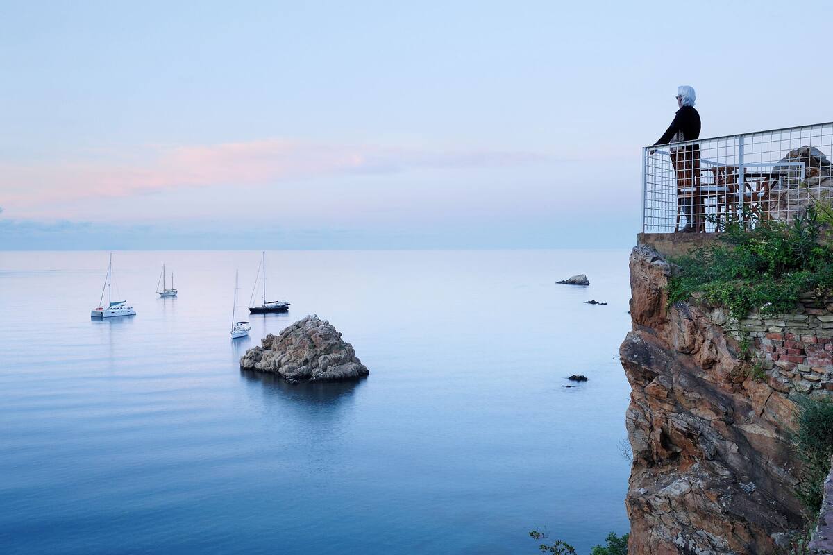 A coastal scene with a rocky cliff and a person standing on a balcony overlooking the sea.