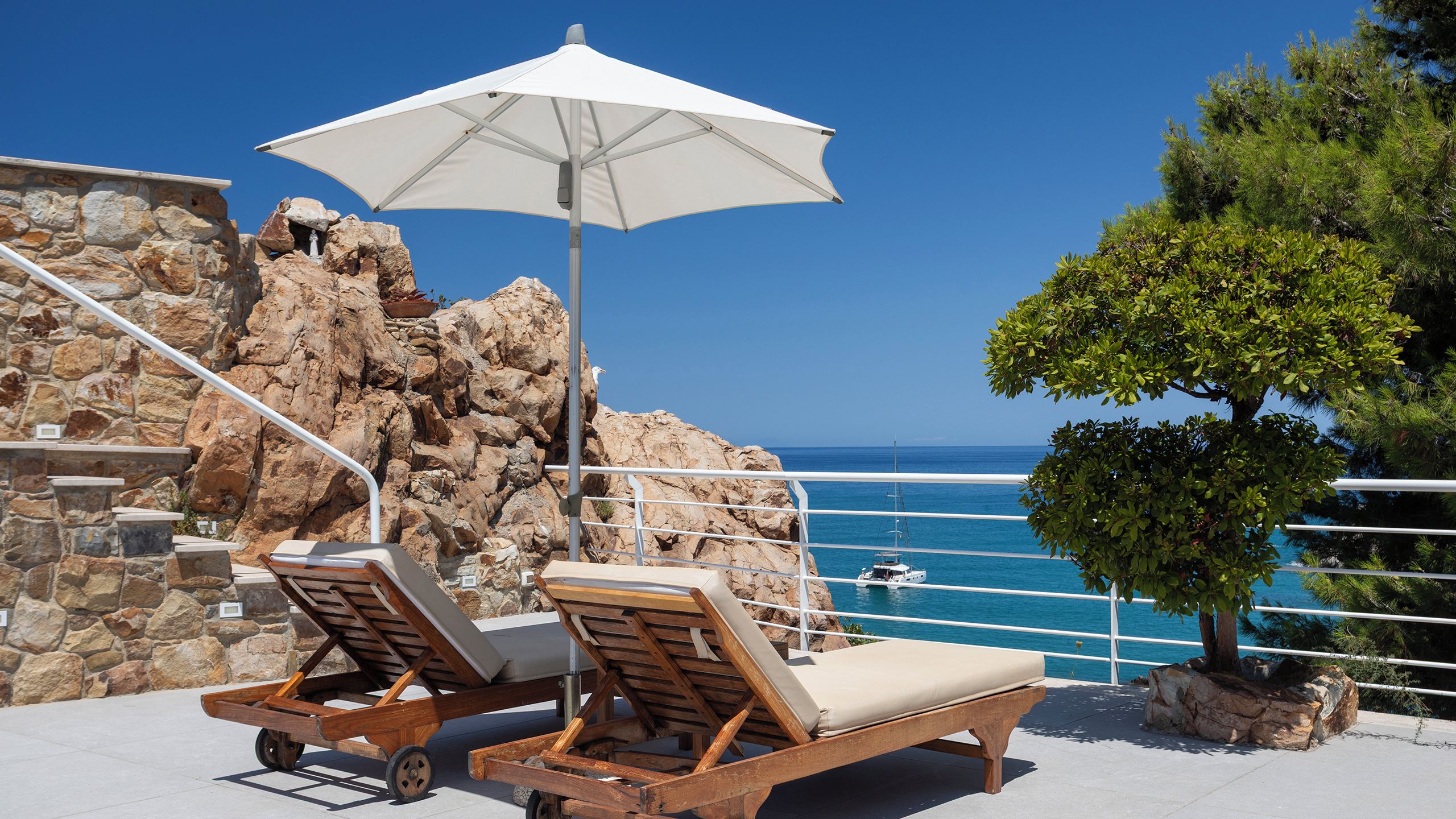 A seaside terrace with two wooden lounge chairs, a white umbrella, and a view of the ocean.