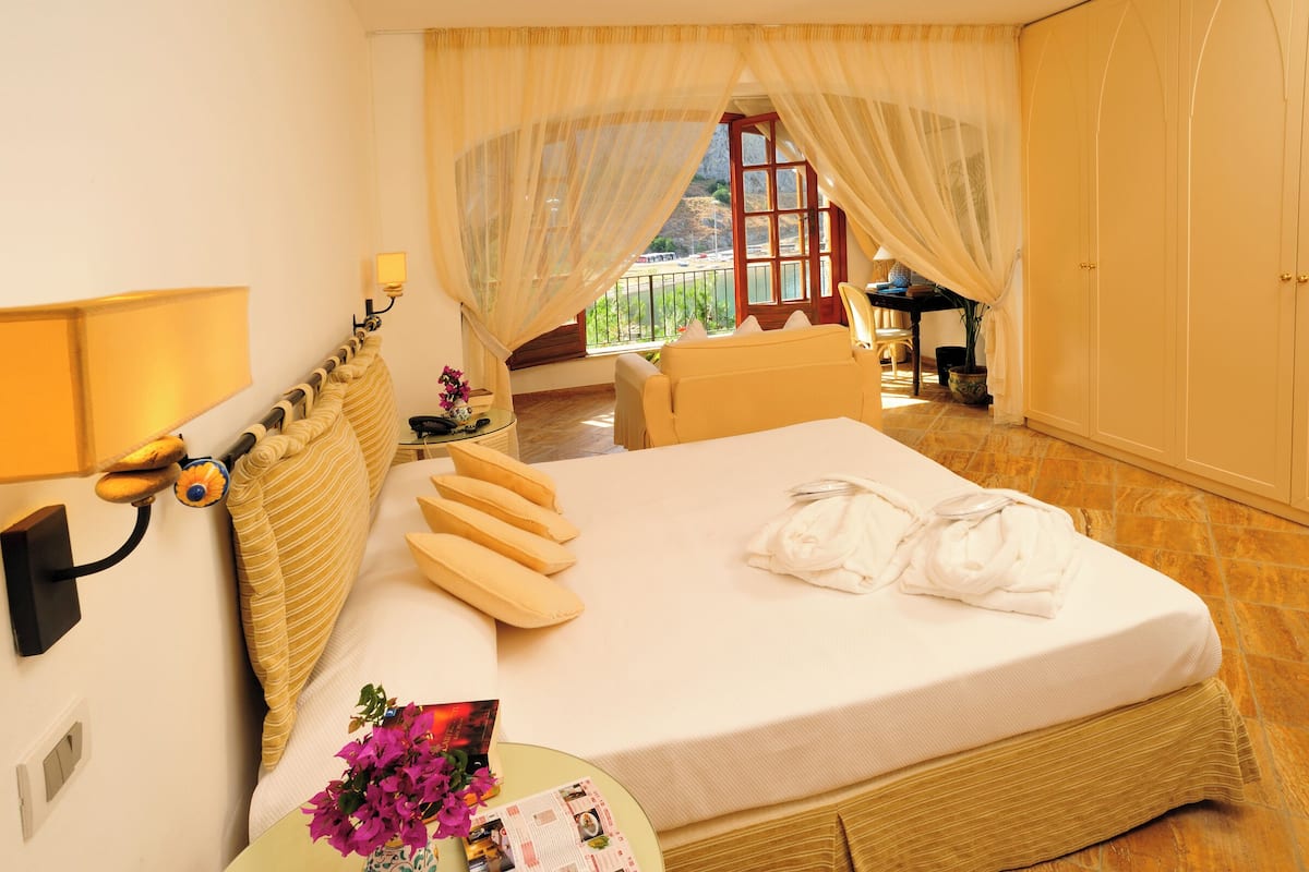 A hotel room with a bed, a nightstand, a window with curtains, a table with a lamp, and a view of the outside.