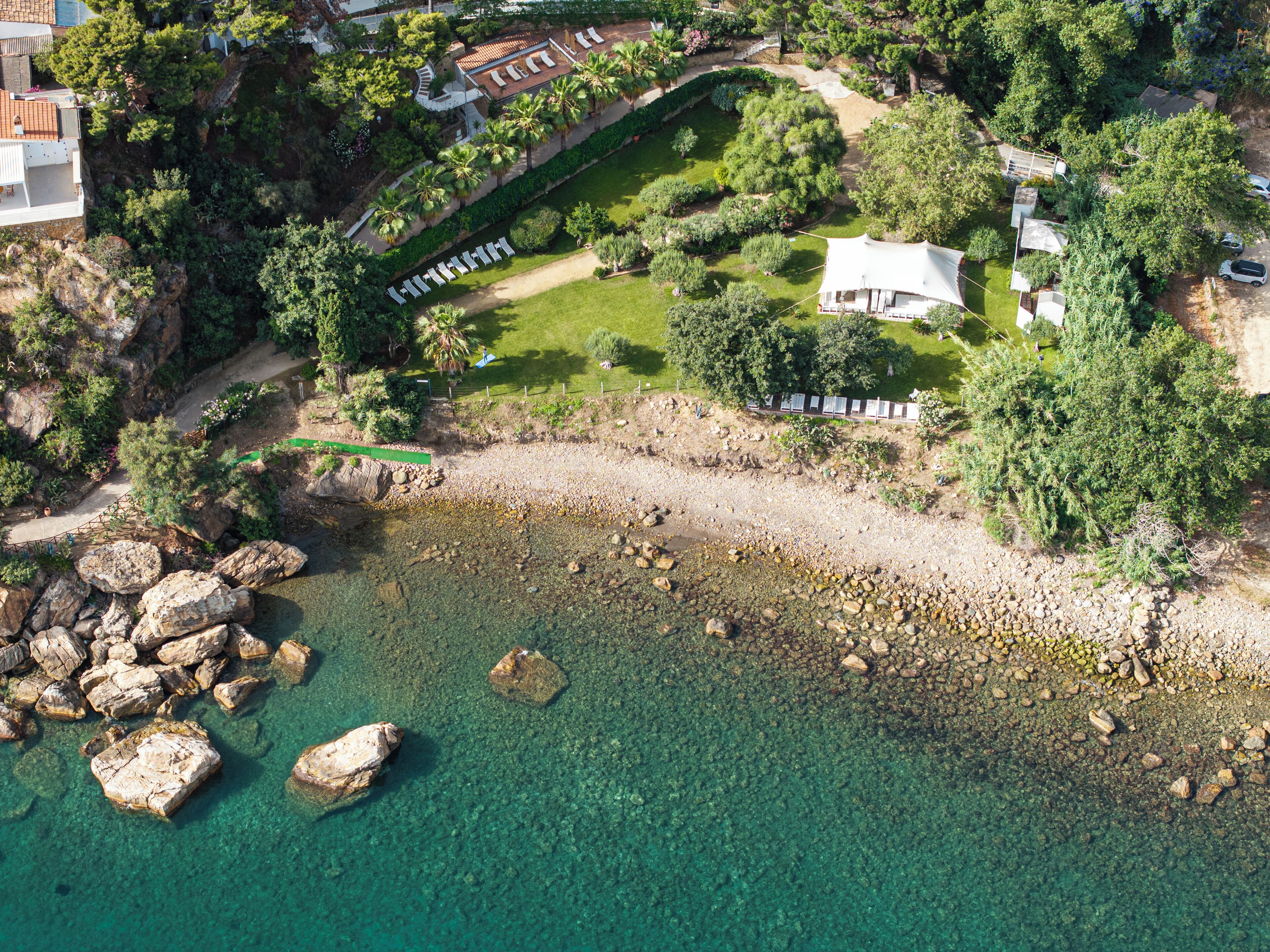 Vista aérea de una propiedad costera con playa despejada, costa rocosa y abundante vegetación.