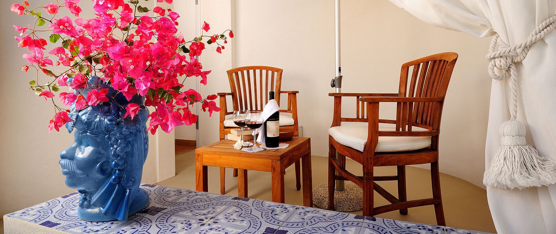 A room with a blue vase of pink flowers, two wooden chairs, a small table with a bottle and glasses, and a patterned tablecloth.