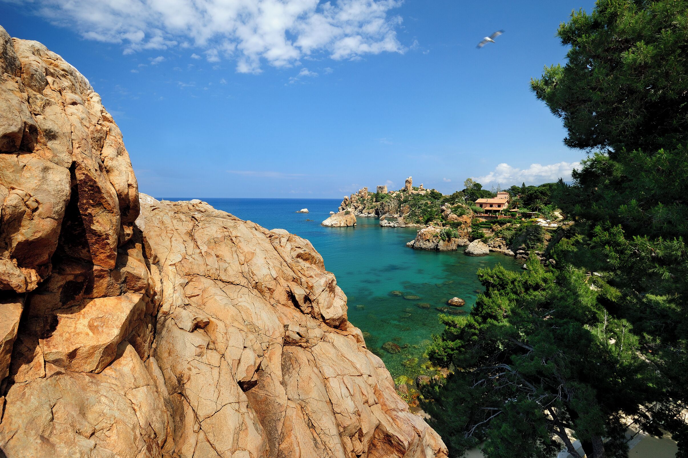 A coastal landscape with rocky cliffs, clear blue sea, and a small building on a hill.