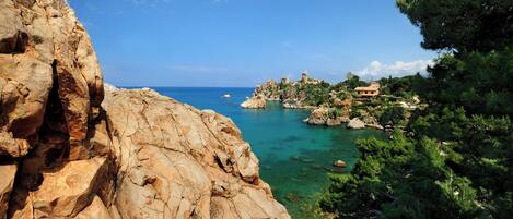 A coastal landscape with rocky cliffs, clear blue sea, and a small building on a hill.