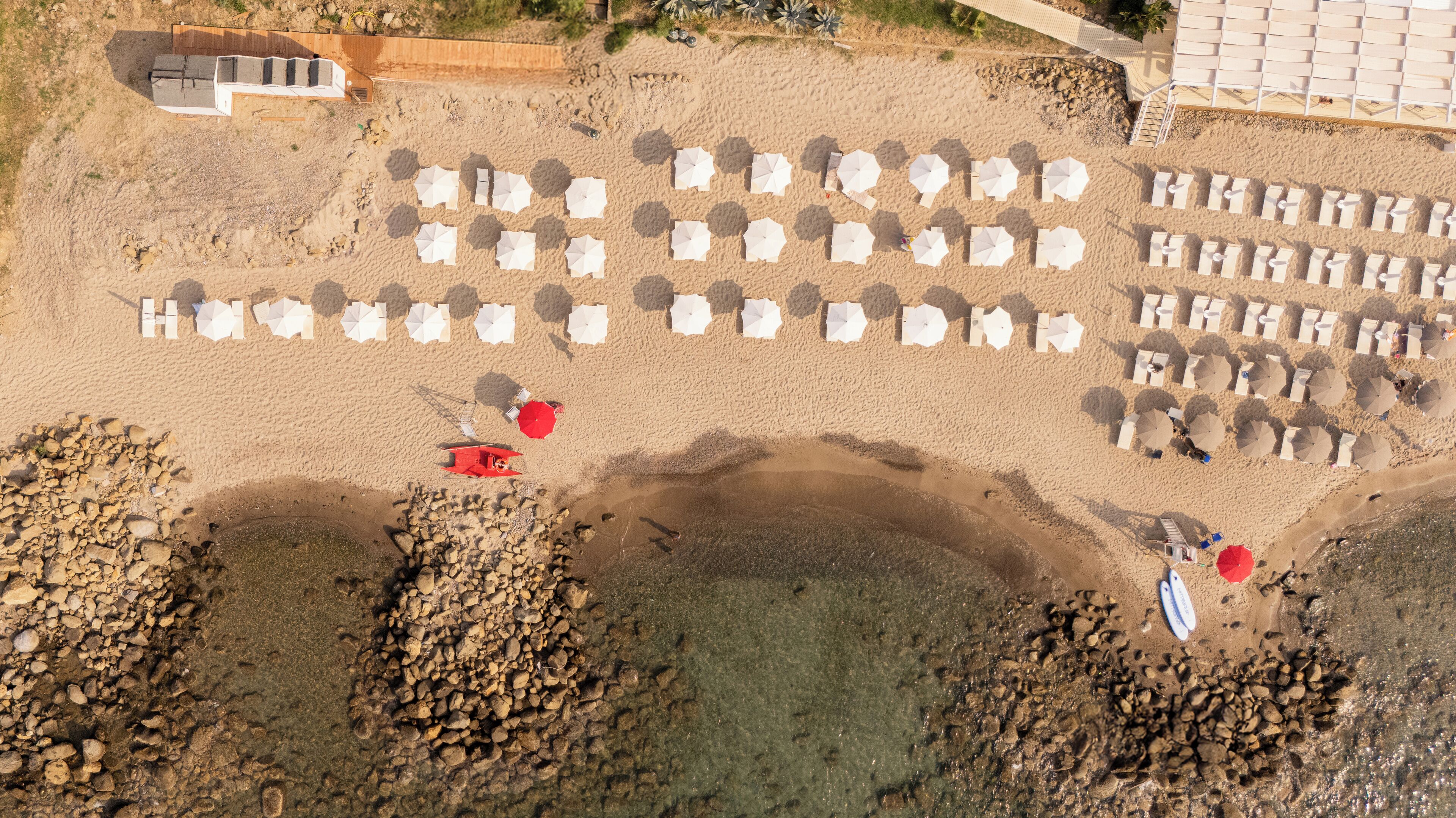 An aerial view of a beach with rows of white umbrellas and beach chairs, a red boat, and rocky areas near the water.