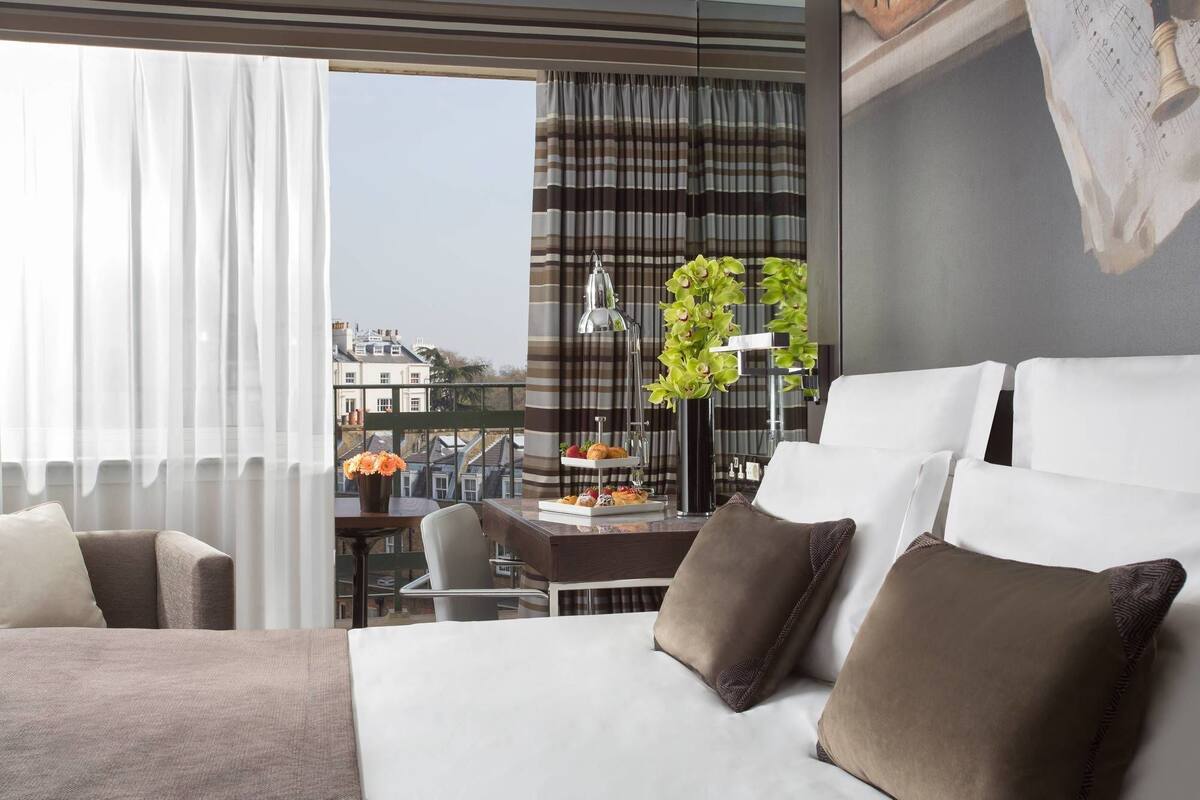 Ein modernes Hotelzimmer mit einem großen Bett, einem Esstisch mit Blumenvase, einer Lampe und Blick auf die Stadtlandschaft.