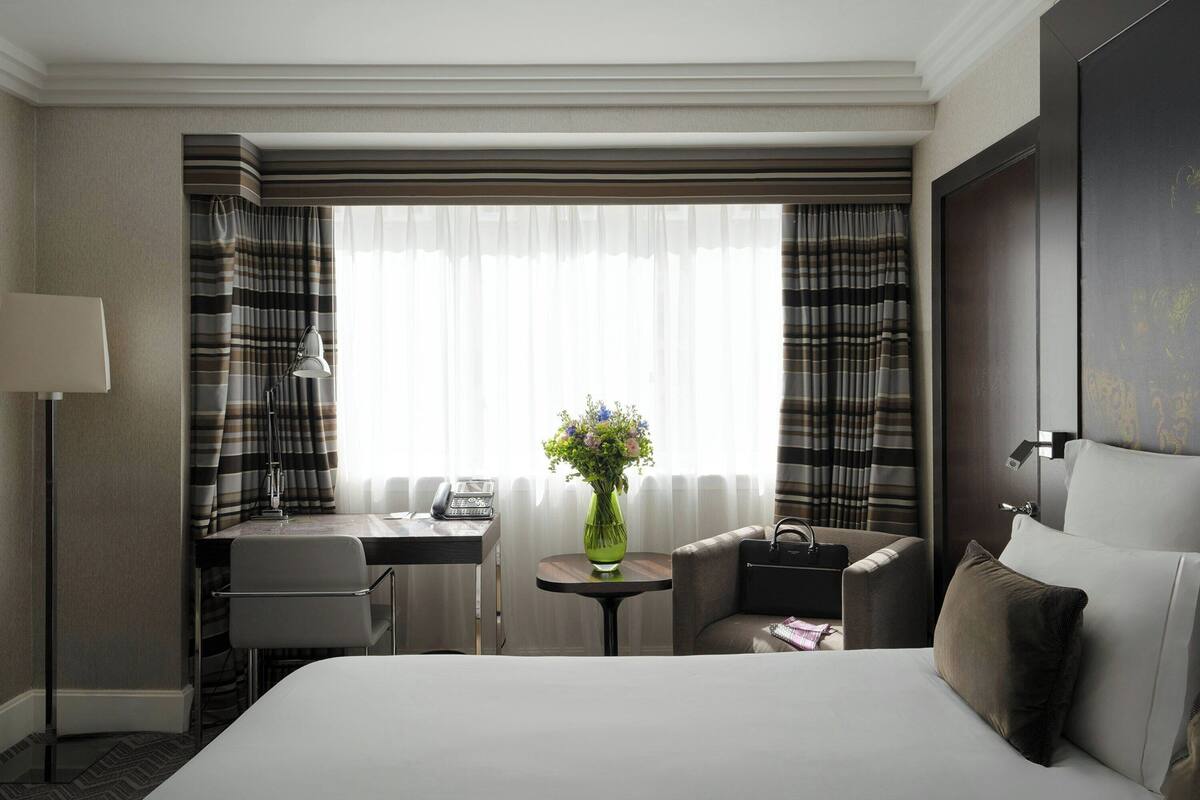 Ein Hotelzimmer mit einem Bett, einem Schreibtisch, einem Stuhl, einem kleinen Tisch mit Blumenvase und einem Vorhangfenster.