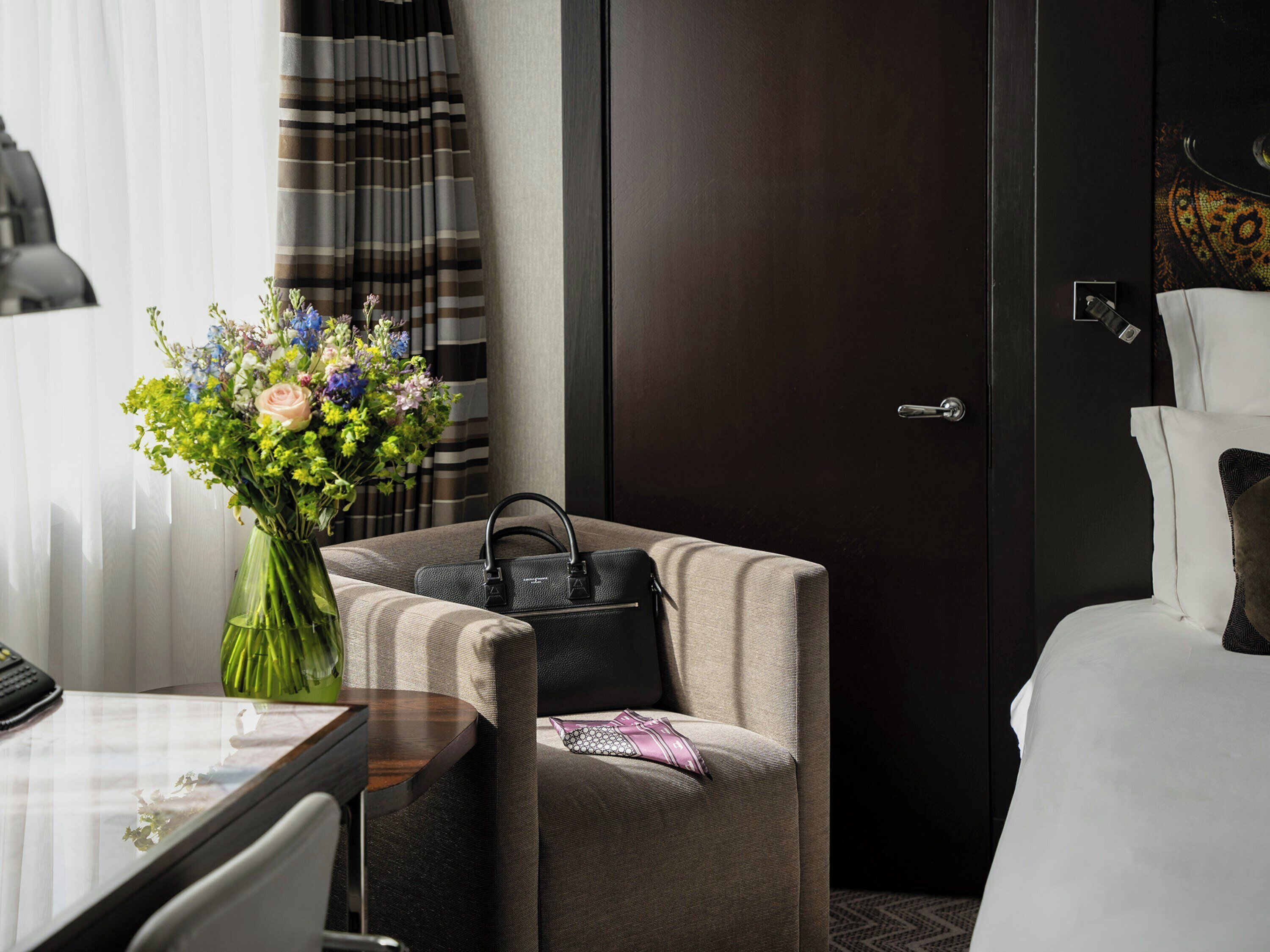 A hotel room with a bed, a chair, a desk, a vase with flowers, and a door.