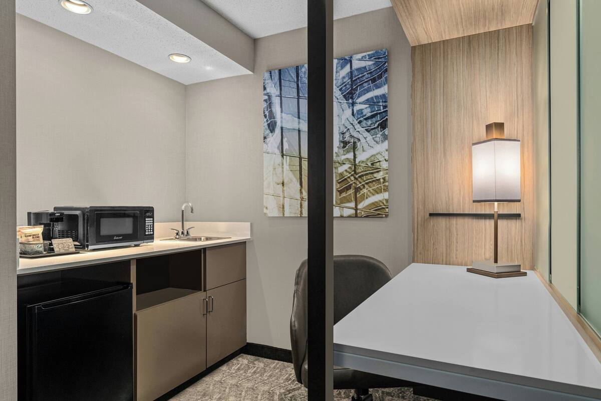 A hotel room with a kitchenette, a desk, a chair, a lamp, and artwork on the walls.