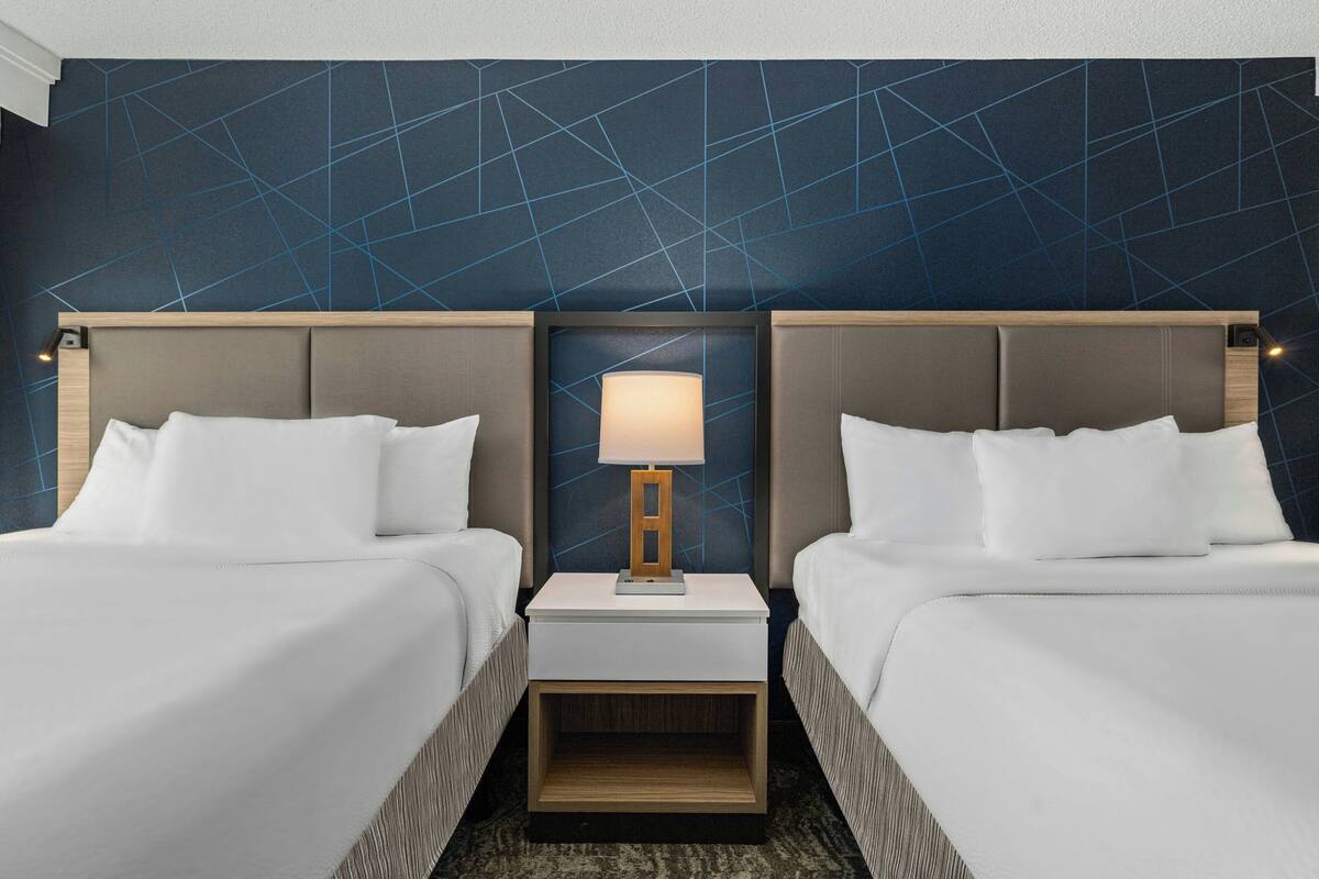 A hotel room with two beds, a nightstand with a lamp, and a geometric-patterned wall.