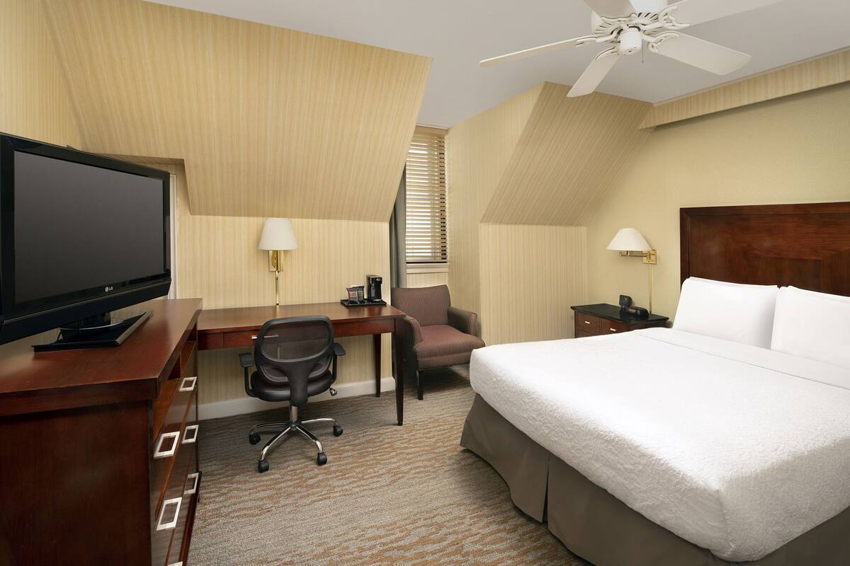 A hotel room with a bed, desk, chair, television, lamps, and a ceiling fan.