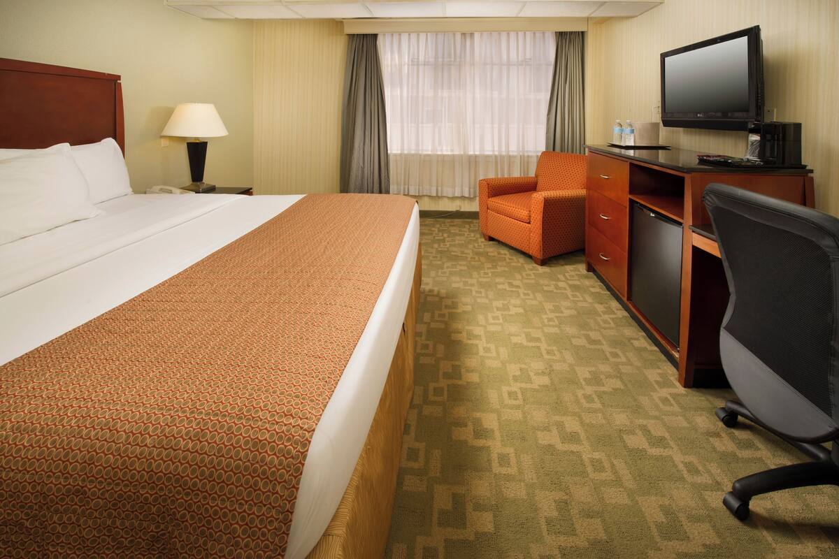 A hotel room with a large bed, a desk with a television, a chair, and a desk lamp.