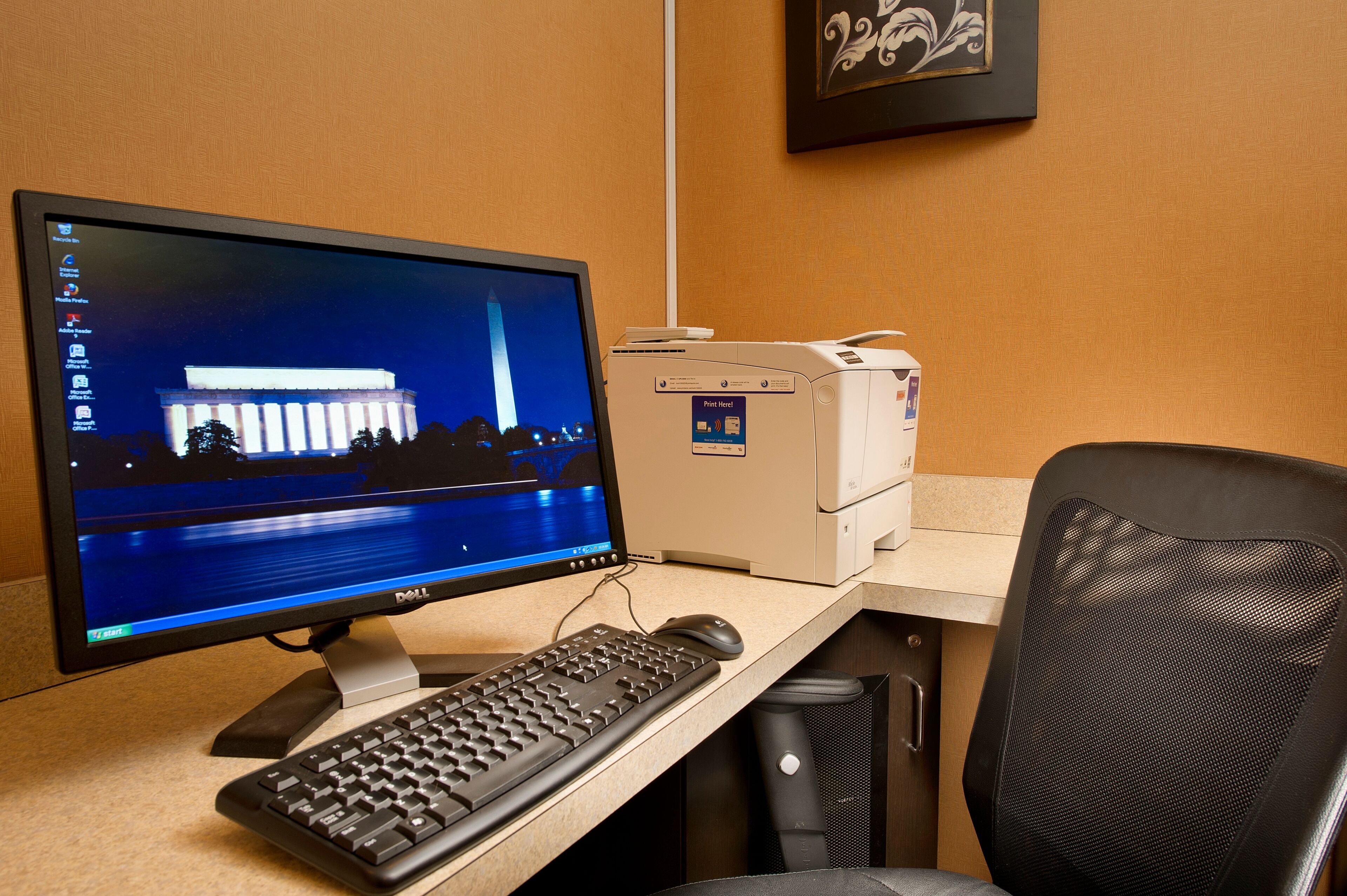 Una computadora de escritorio con un monitor mostrando una escena nocturna del Monumento a Washington, un teclado, un ratón y una impresora sobre un escritorio.