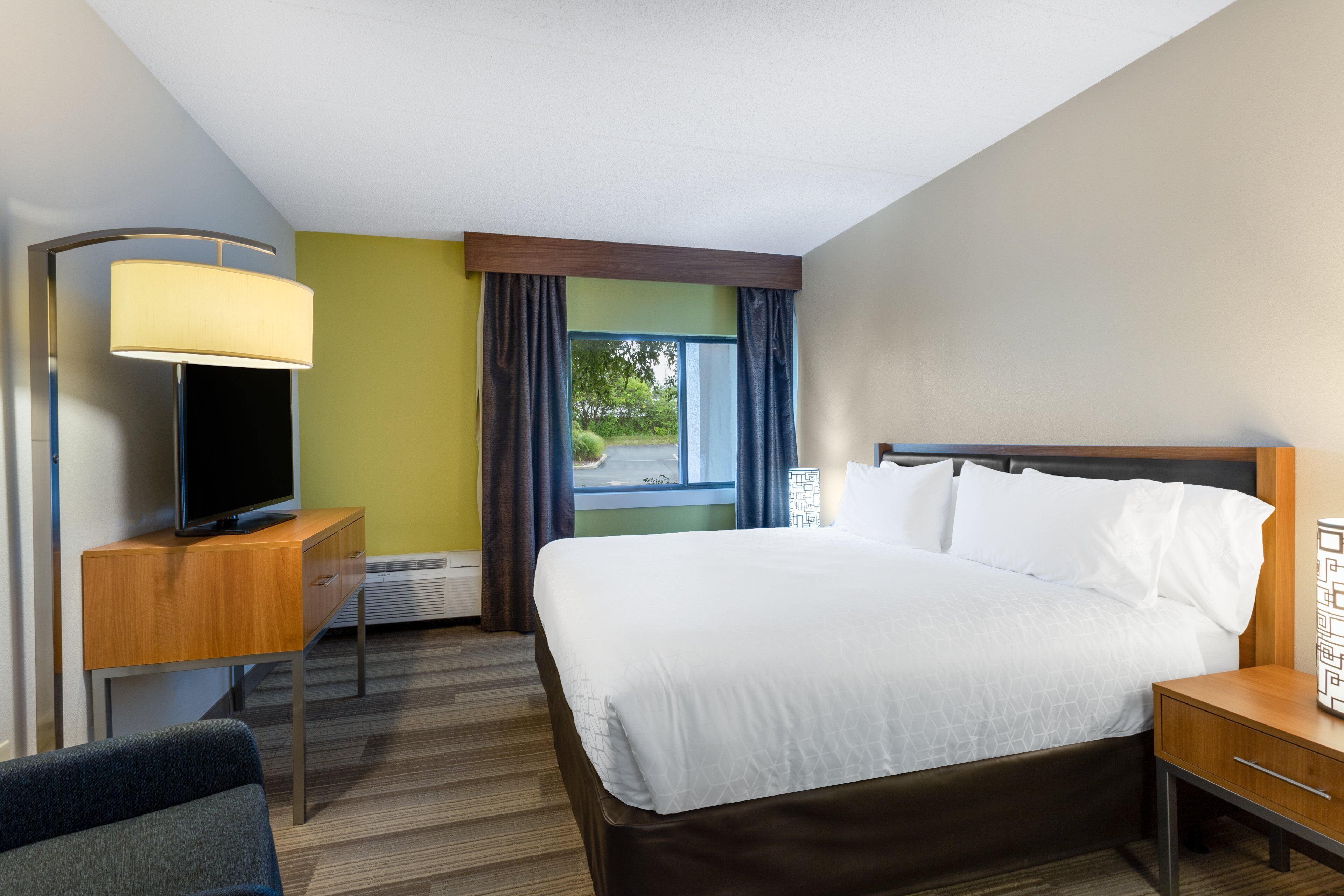 A hotel room with a bed, a desk with a lamp, a chair, and a window with curtains.