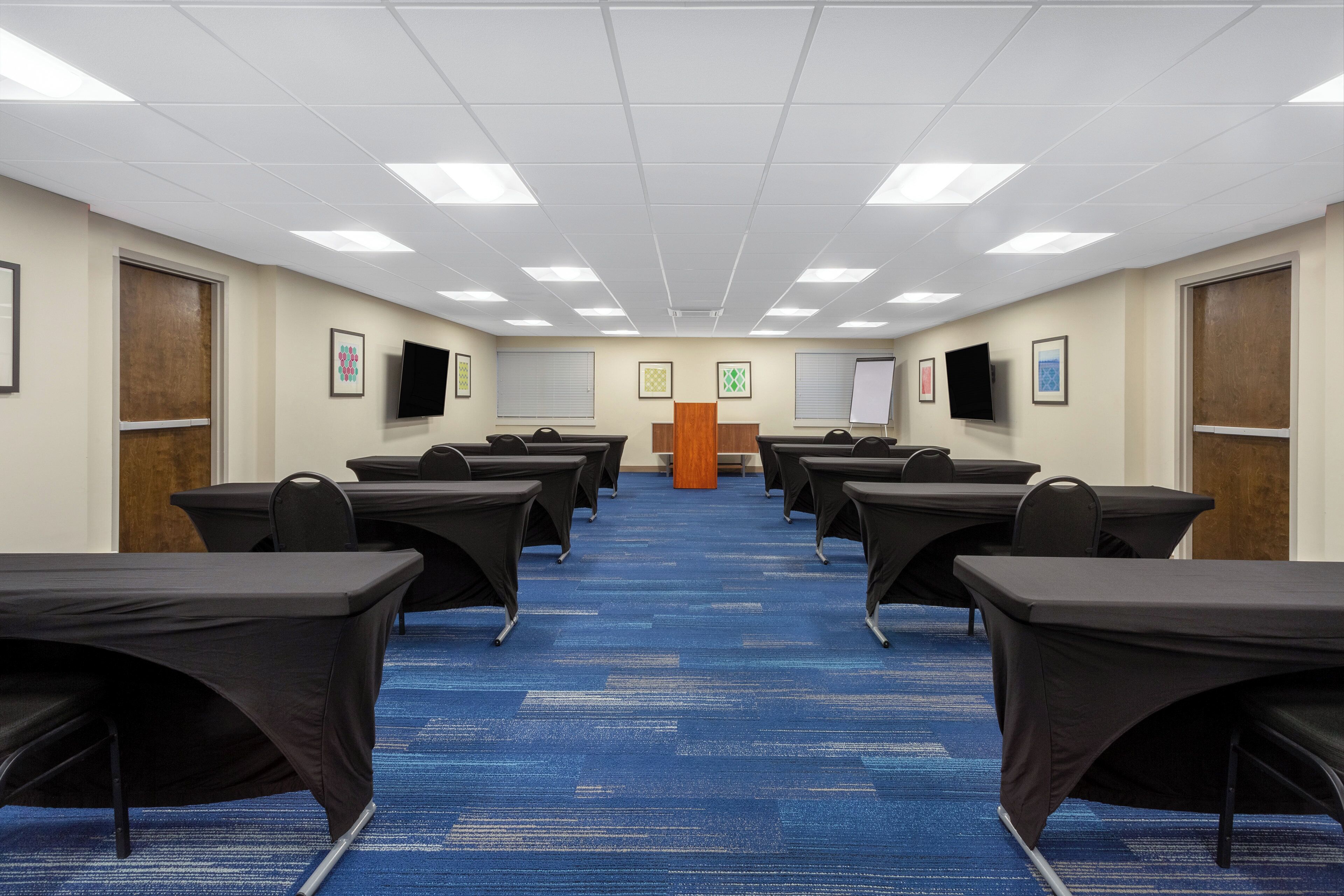 A conference room with rows of tables and chairs, a podium, and framed pictures on the walls.