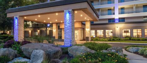 A hotel entrance with a covered outdoor area, stone pillars, and a landscaped garden with rocks and plants.
