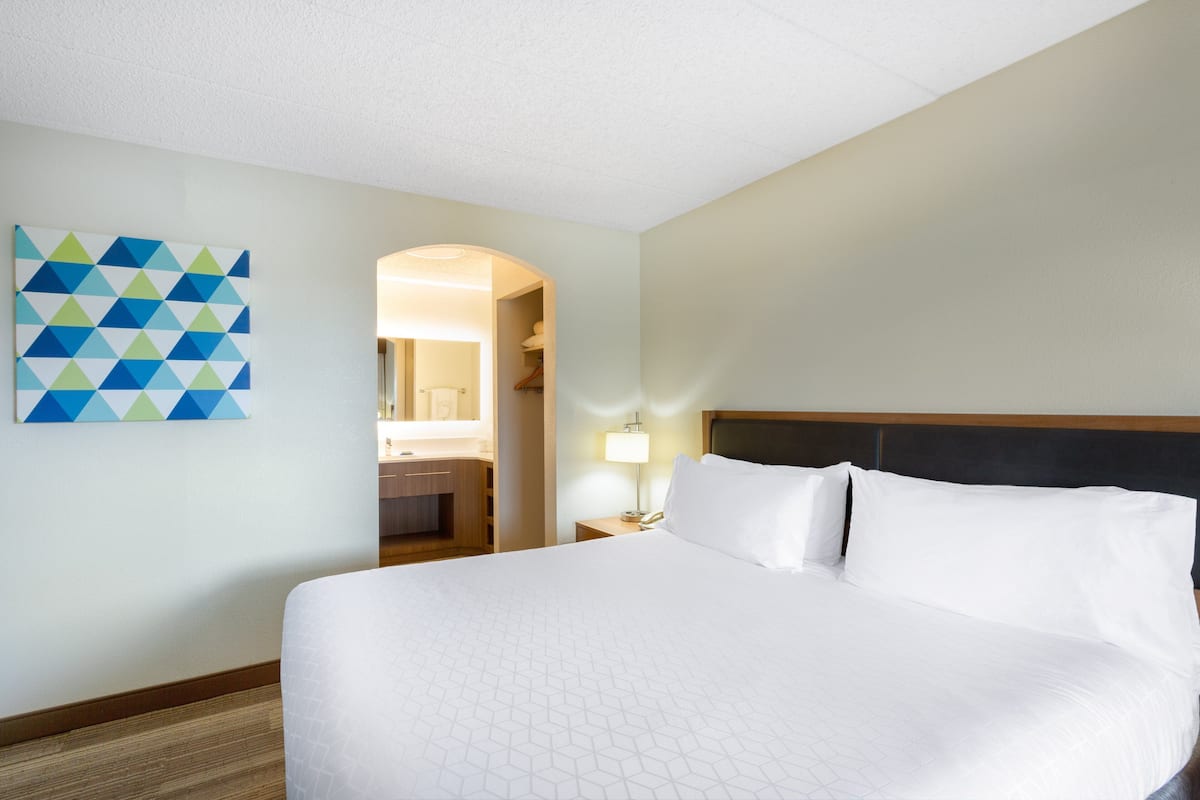 A hotel room with a large bed, a wall-mounted geometric artwork, a nightstand with a lamp, and a view into a bathroom.