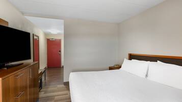 Une chambre d’hôtel avec un lit, un téléviseur posé sur une commode en bois, une table de chevet avec une lampe, et une porte rouge en arrière-plan.